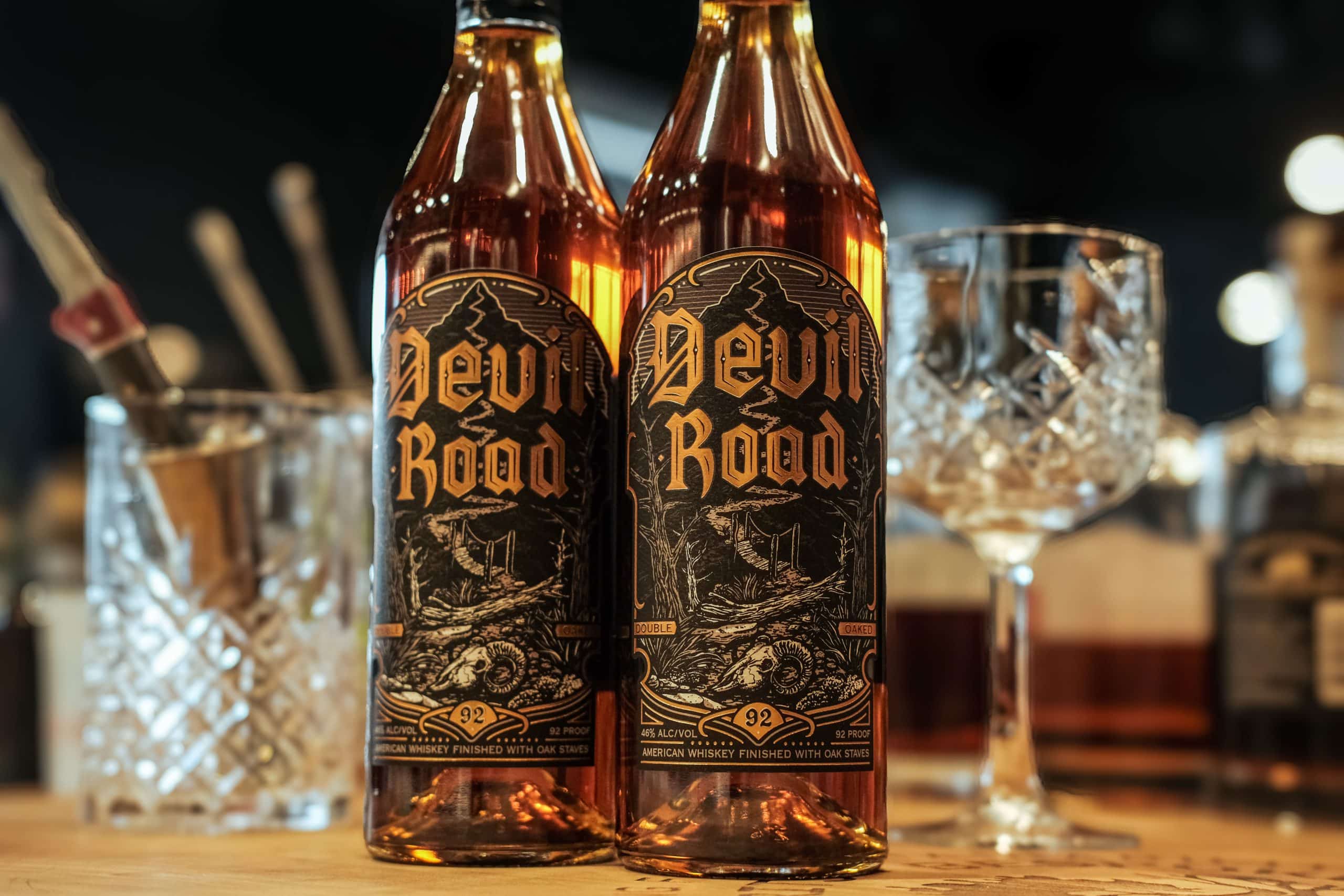 Two bottles of Devil Road American Whiskey with fancy cocktail glasses and bartending tools