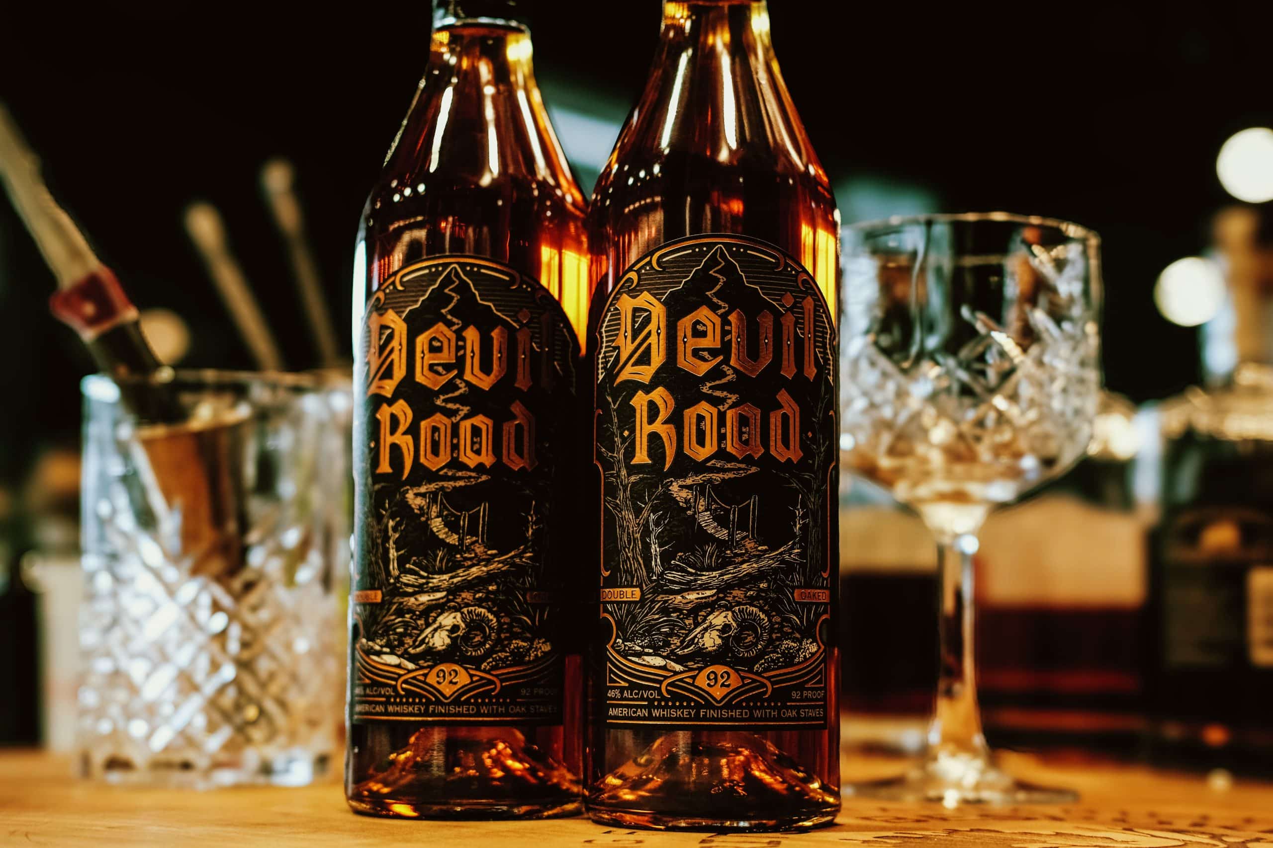 Two bottles of Devil Road American Whiskey with fancy cocktail glasses and bartending tools